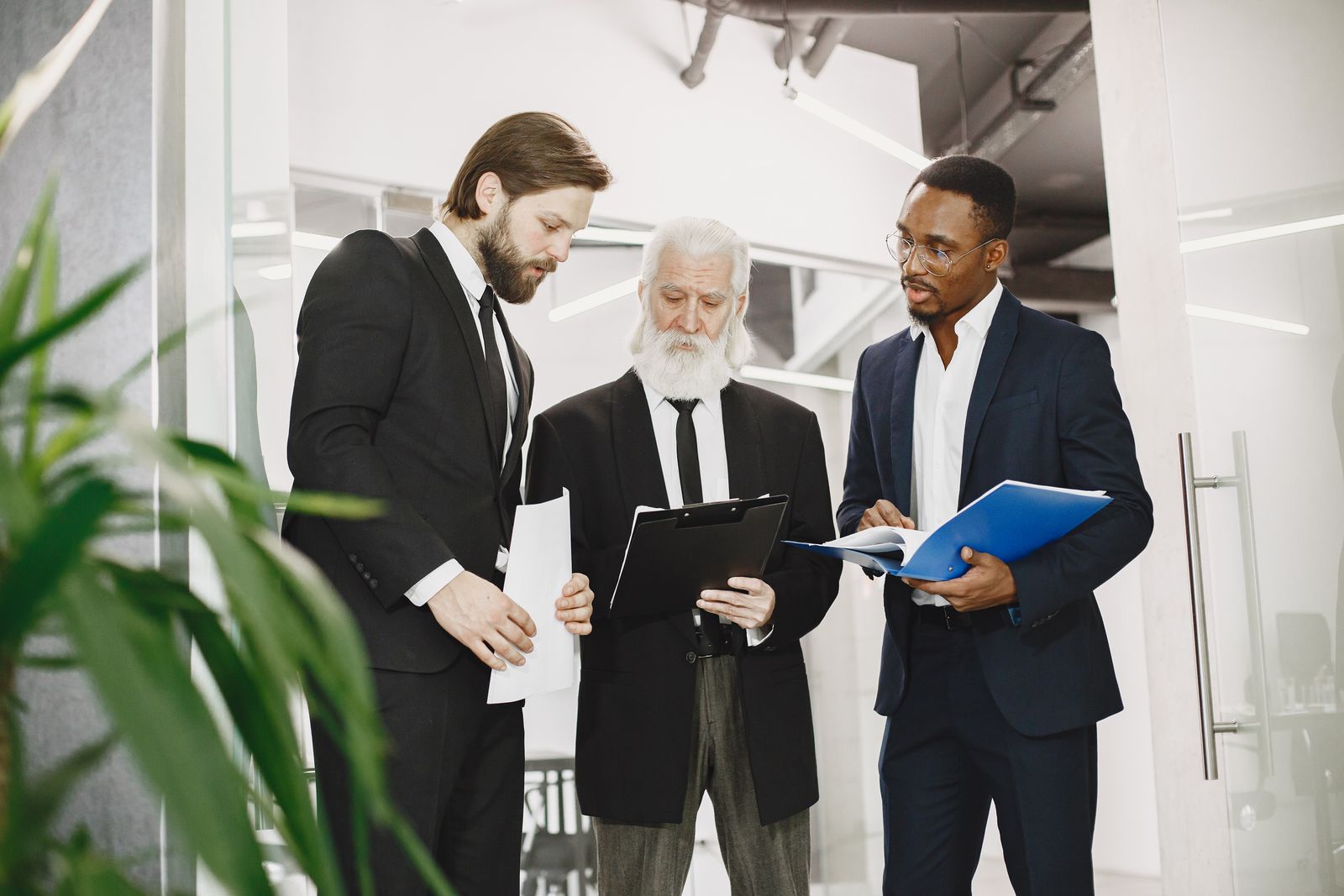Three fund principals in dark suits reviewing loan folders in a modern office corridor